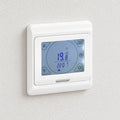 Weißes digitales Wandthermostat mit blauem Touchscreen an einer hellen Putzwand.